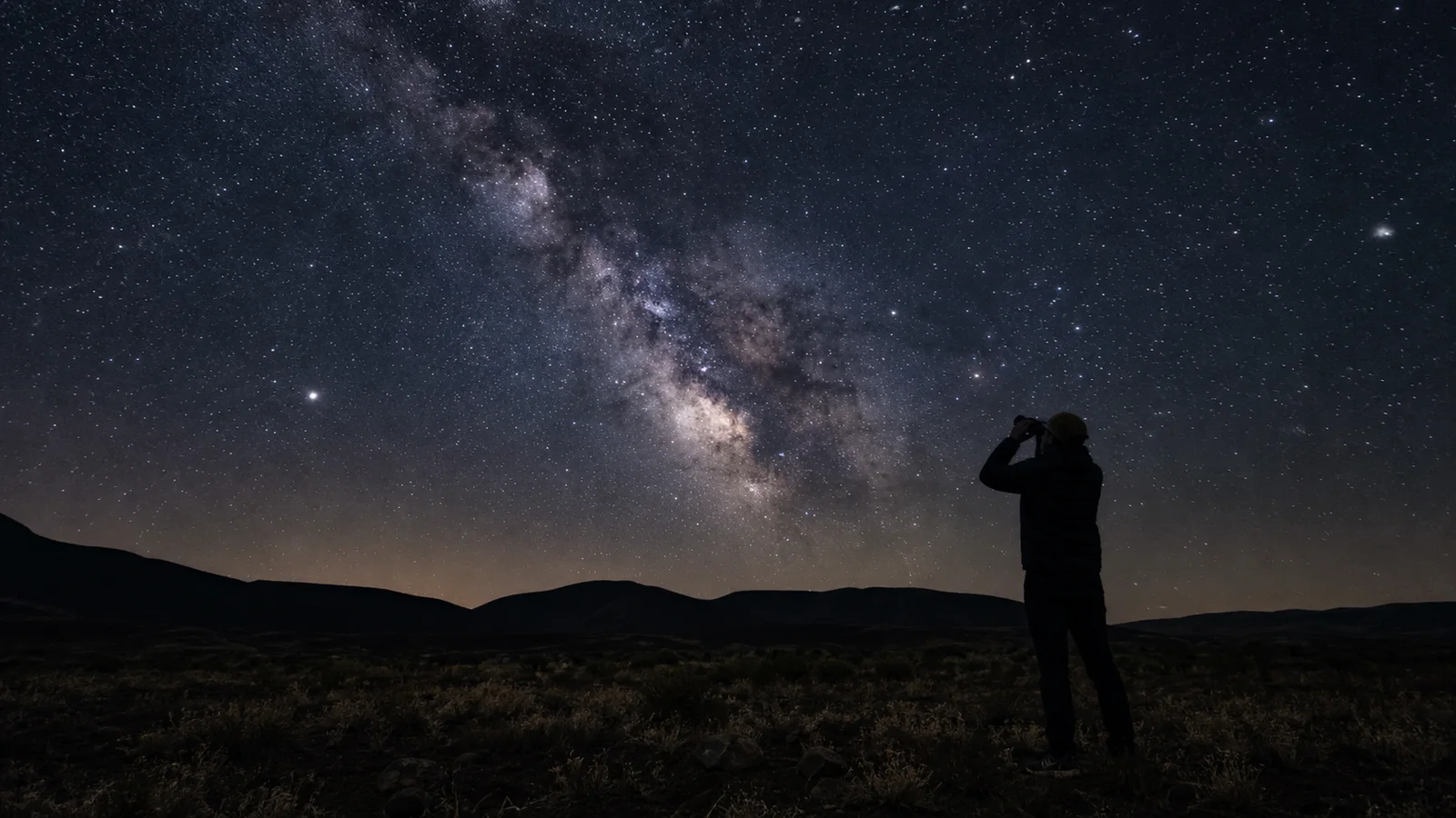 A person uses binoculars under a dark sky filled with the Milky Way.