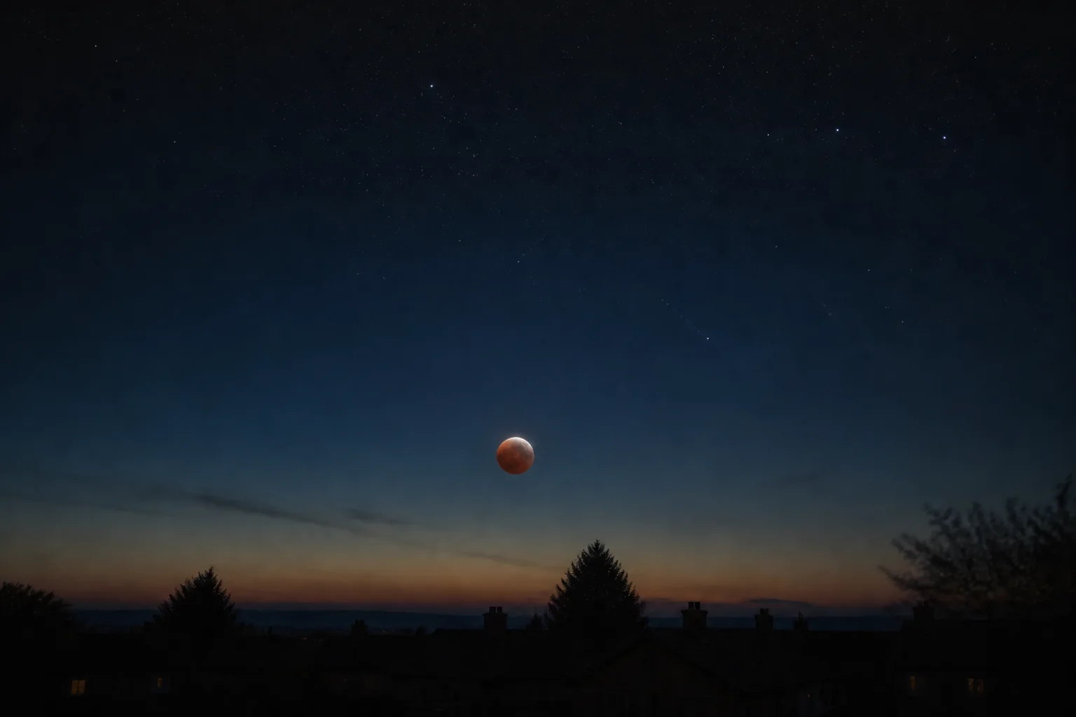 Partial lunar eclipse low above distant rooflines and trees near dawn