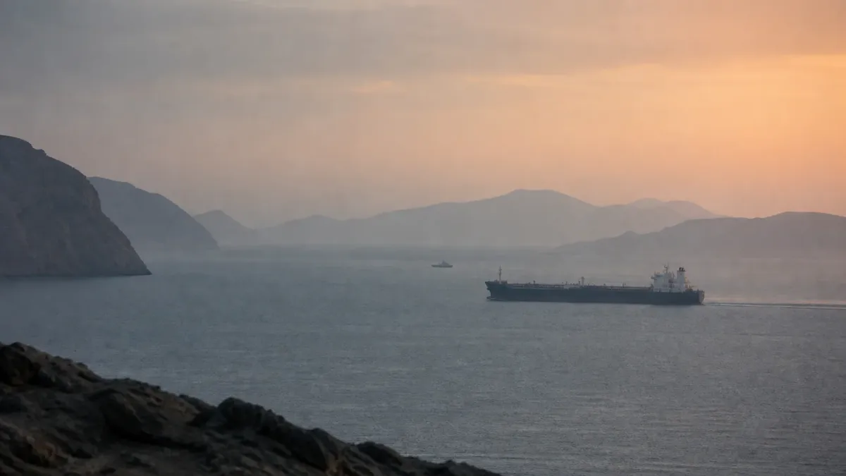 A commercial tanker crosses a calm, hazy gulf waterway at dawn as a distant patrol vessel remains on watch.
