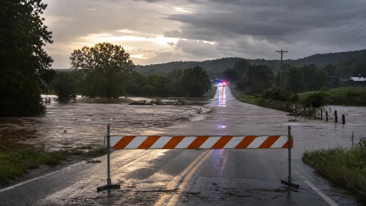 A generic flooded rural road blocked by a barricade with emergency lights in the distance
