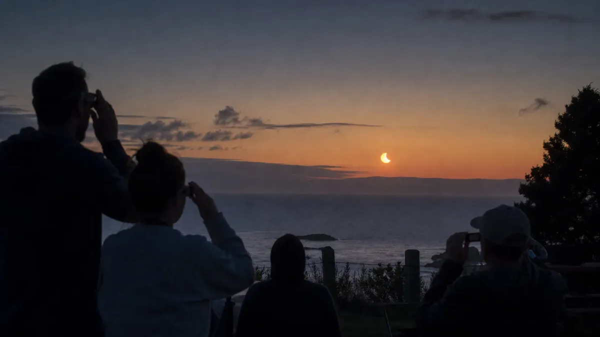 People wearing eclipse glasses watch a partial solar eclipse near a coastal horizon at dusk