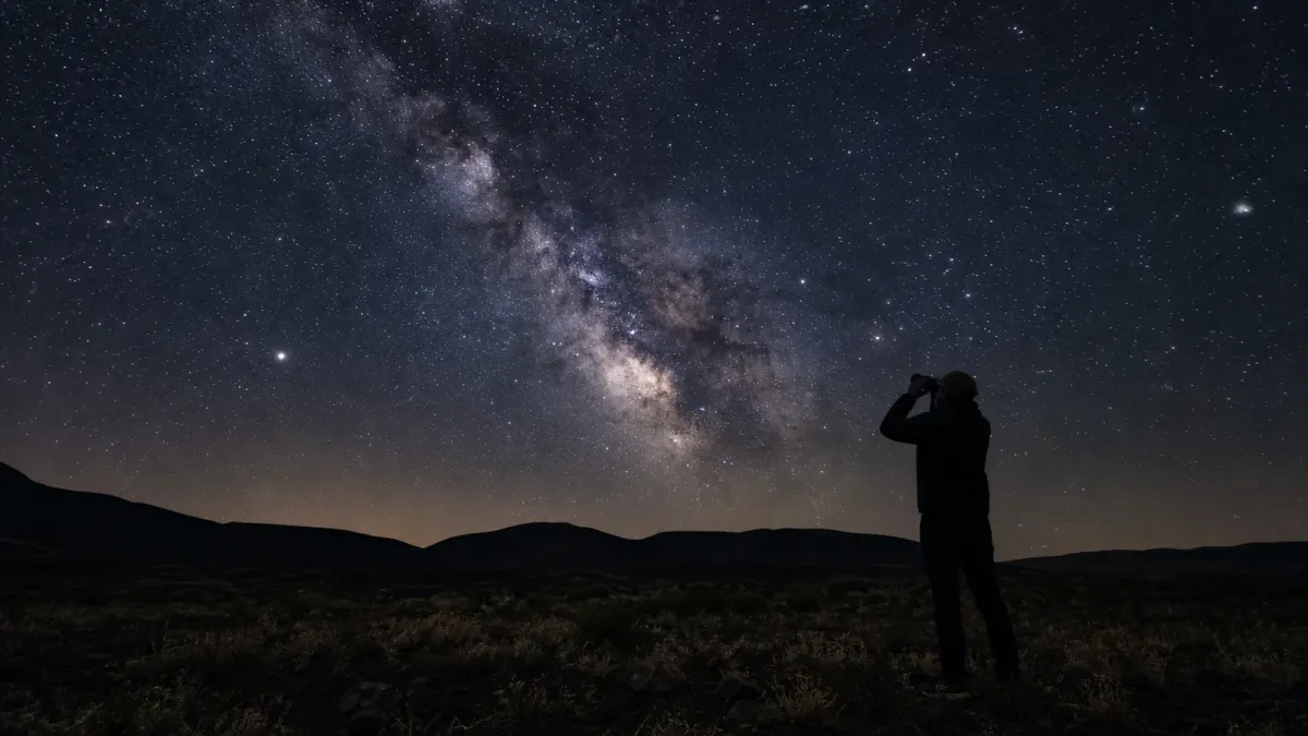 A person uses binoculars under a dark sky filled with the Milky Way.