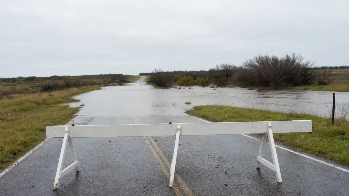 A temporary barricade blocks a rural road where muddy floodwater crosses the pavement.