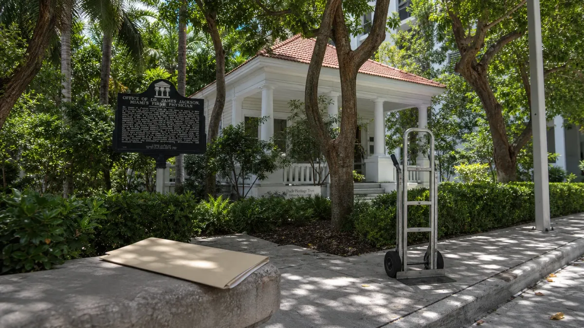 Historic Jackson Cottage and Dade Heritage Trust setting in Brickell framed with a civic folder and moving dolly to suggest the approved sale and possible relocation.