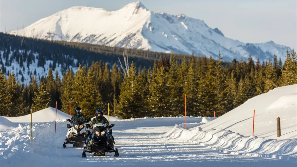 Snowmobilers travel on a groomed winter road in Yellowstone National Park with snowy mountains in the background.