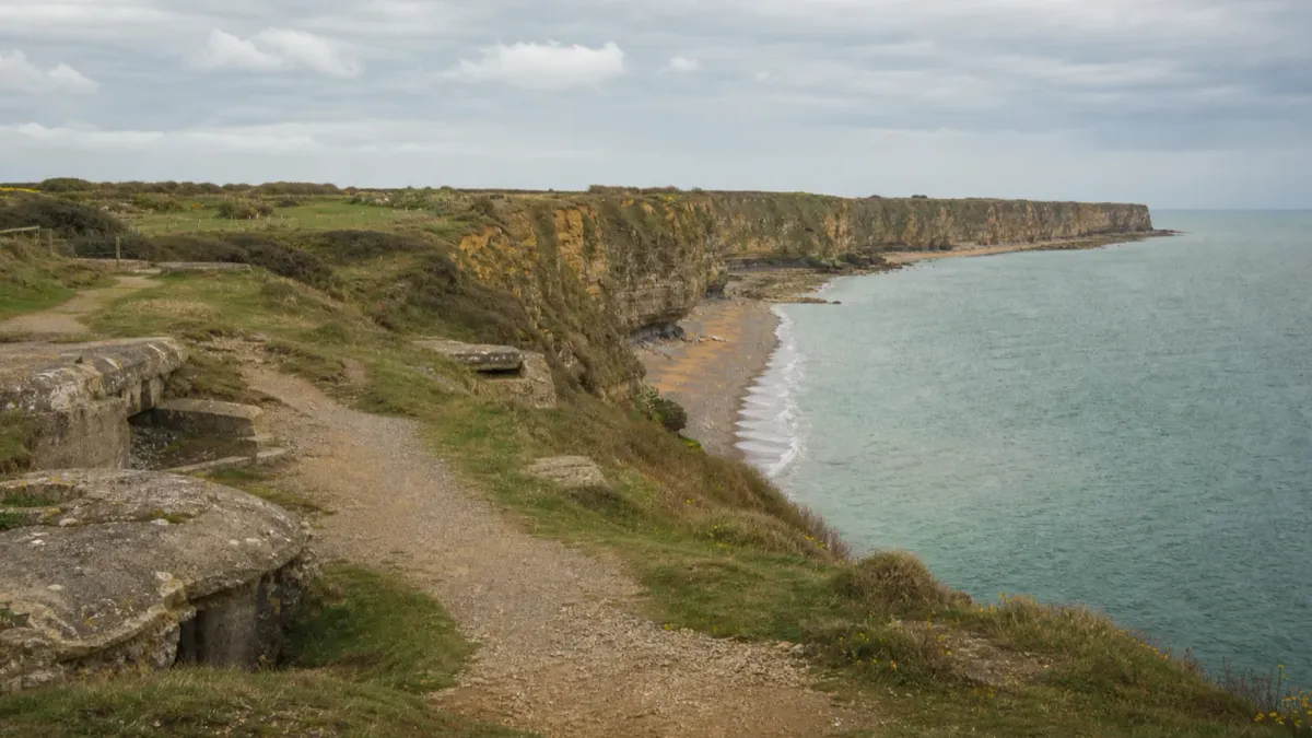 A grounded editorial view of Pointe du Hoc above the Normandy coast with preserved paths and coastal defenses in the foreground
