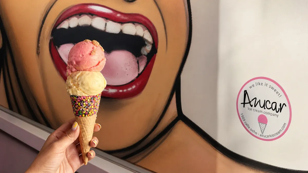 A two-scoop waffle cone held before Azucar Ice Cream's signature painted mouth mural with the shop's circular brand seal nearby