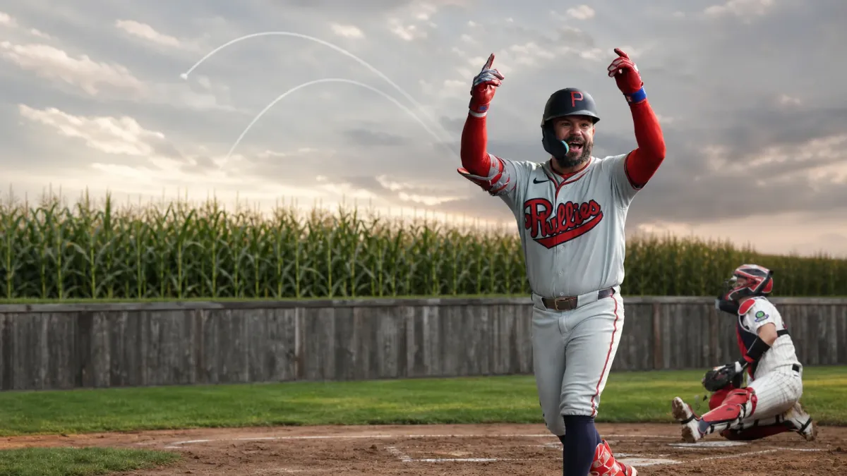 Kyle Schwarber in a gray Phillies retro uniform against the Field of Dreams cornfield with two subtle home-run arc traces