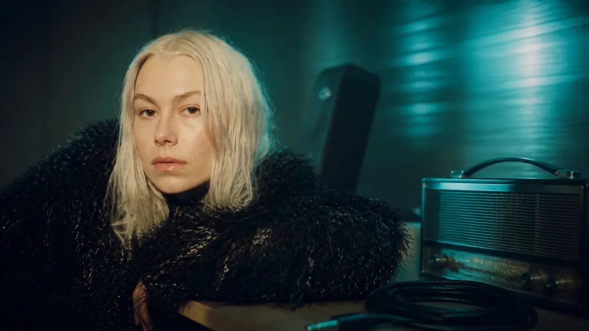 Phoebe Bridgers in a cyan-lit editorial listening room with an analog radio and instrument cable