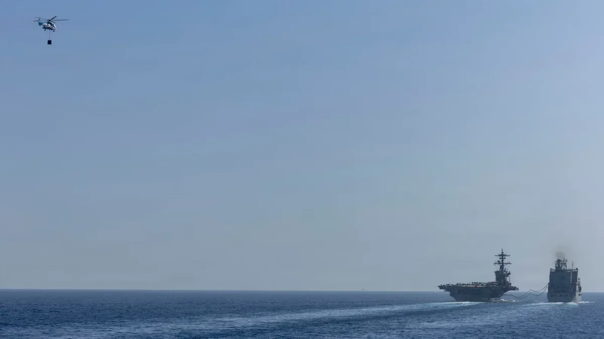 USS Abraham Lincoln sails beside a replenishment oiler as a helicopter carries cargo overhead.