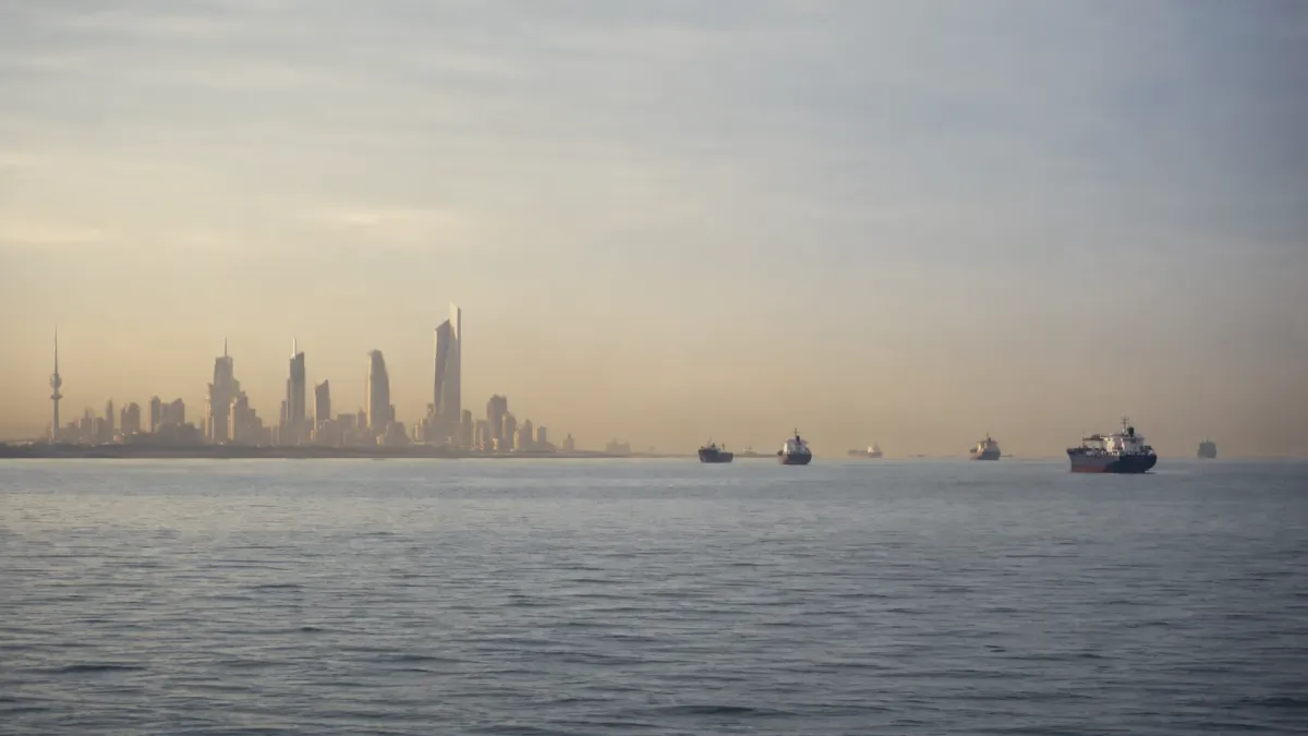 Kuwait's recognizable waterfront skyline across calm Gulf water with commercial vessels in a hazy shipping corridor