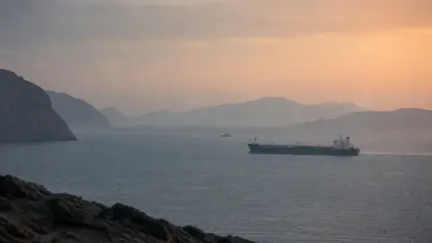 A commercial tanker crosses a calm, hazy gulf waterway at dawn as a distant patrol vessel remains on watch.