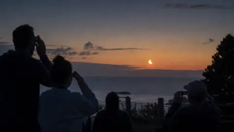 People wearing eclipse glasses watch a partial solar eclipse near a coastal horizon at dusk