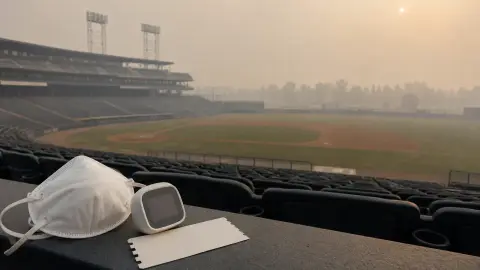 A hazy baseball field viewed from empty stands with a mask, blank ticket and small air-quality monitor in the foreground.