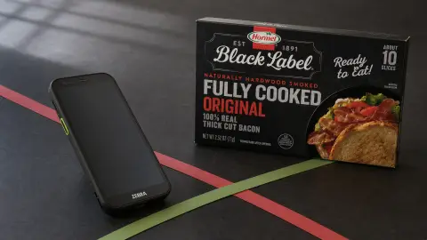 A Zebra mobile computer positioned beside a Hormel Black Label package as red and green paper lines cross on a dark work surface.