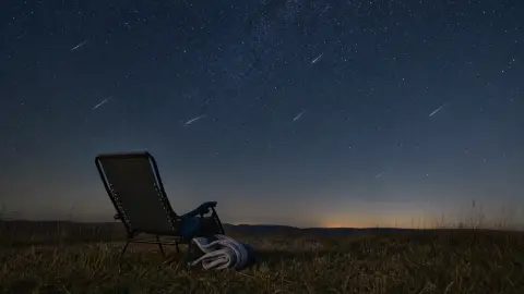 A dark rural viewing spot with a reclining chair beneath subtle meteor streaks in a wide night sky.