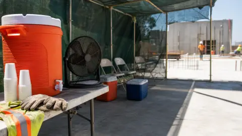 A shaded worksite break area with water, cups, gloves and a fan set apart from sunlit concrete.