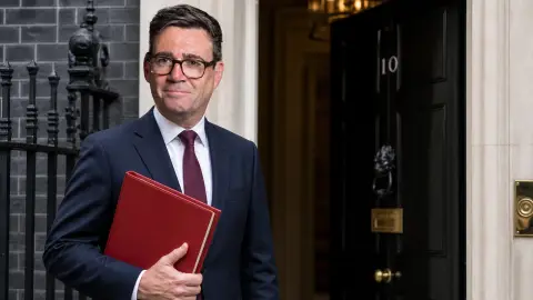 Andy Burnham shown beside the black door of 10 Downing Street with a red ministerial folder, illustrating Britain's leadership handover