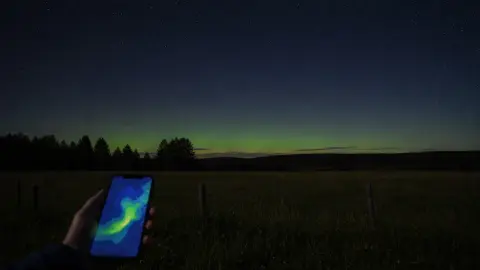 A quiet northern horizon with faint green aurora light above a dark field as a phone checks a live forecast map