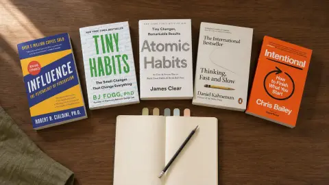 Influence, Tiny Habits, Atomic Habits, Thinking, Fast and Slow and Intentional arranged around a blank notebook on a warm reading desk