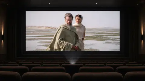 A wide editorial composite of Matt Damon as Odysseus and Zendaya as Athena from The Odyssey shown on a large cinema screen above dark theater seats.