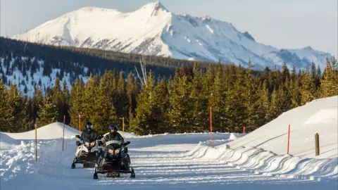 Snowmobilers travel on a groomed winter road in Yellowstone National Park with snowy mountains in the background.