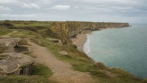 A grounded editorial view of Pointe du Hoc above the Normandy coast with preserved paths and coastal defenses in the foreground