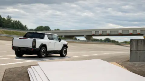 A General Motors vehicle on a Milford Proving Ground-style test lane with blank import paperwork in the foreground, suggesting tariff pressure on auto supply chains.