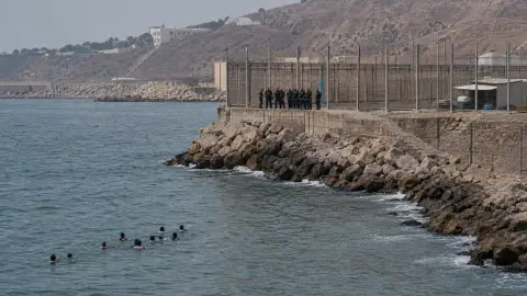Source-grounded editorial view of swimmers approaching Ceuta’s Tarajal breakwater as responders gather above the rocks