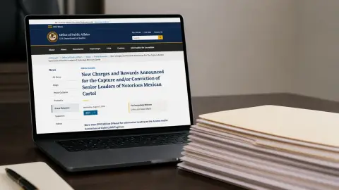 A laptop displays a U.S. Department of Justice press release beside a stack of unmarked federal paperwork.
