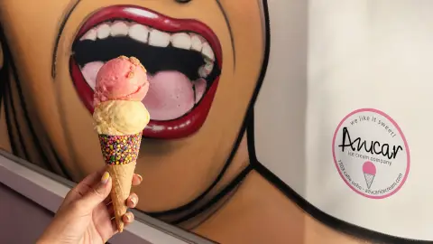 A two-scoop waffle cone held before Azucar Ice Cream's signature painted mouth mural with the shop's circular brand seal nearby
