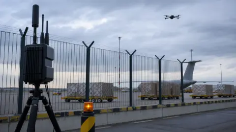 Counter-drone equipment stands beside a fenced cargo-airport perimeter as a small unmarked drone hovers in the distance.