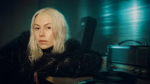 Phoebe Bridgers in a cyan-lit editorial listening room with an analog radio and instrument cable