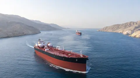 Two intact crude tankers transit a narrow blue-green channel between arid coastlines in a source-grounded editorial view of the Strait of Hormuz.
