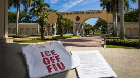 A staged FIU campus threshold with a folded ICE OFF FIU shirt and Regulation 110 packet at the boundary between sunlight and indoor shade.
