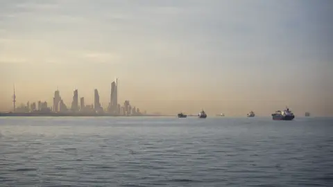 Kuwait's recognizable waterfront skyline across calm Gulf water with commercial vessels in a hazy shipping corridor
